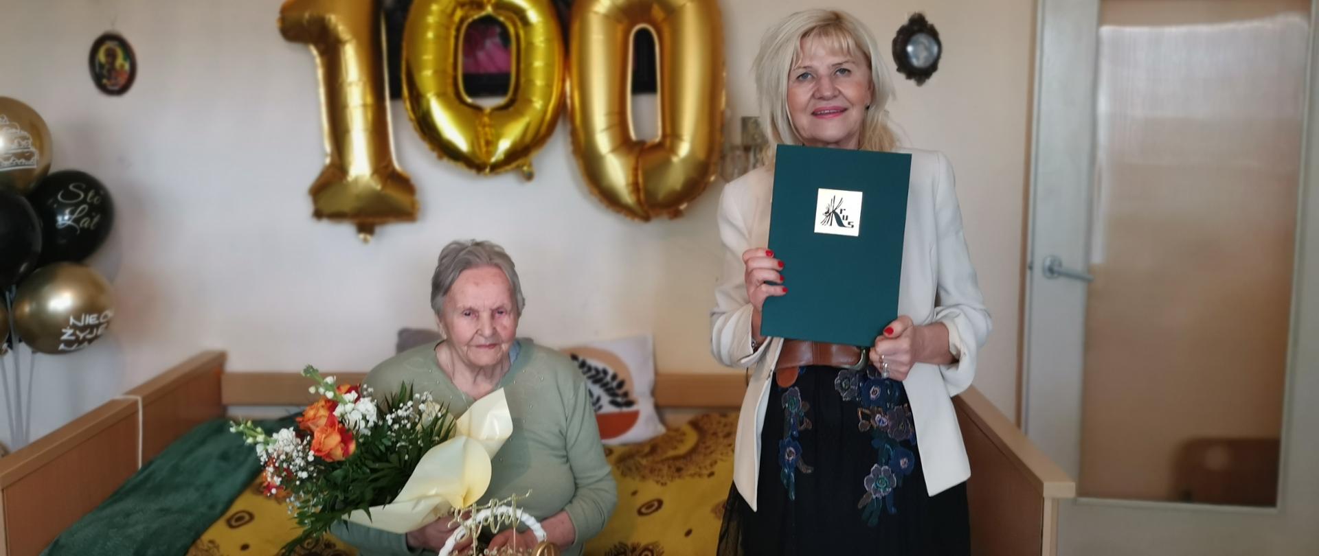Po lewej Jubilatka z bukietem kwiatów siedzi przy stole obok stoi kobieta z dyplomem KRUS w dłoniach., z tyłu balony kształcie liczby 100.