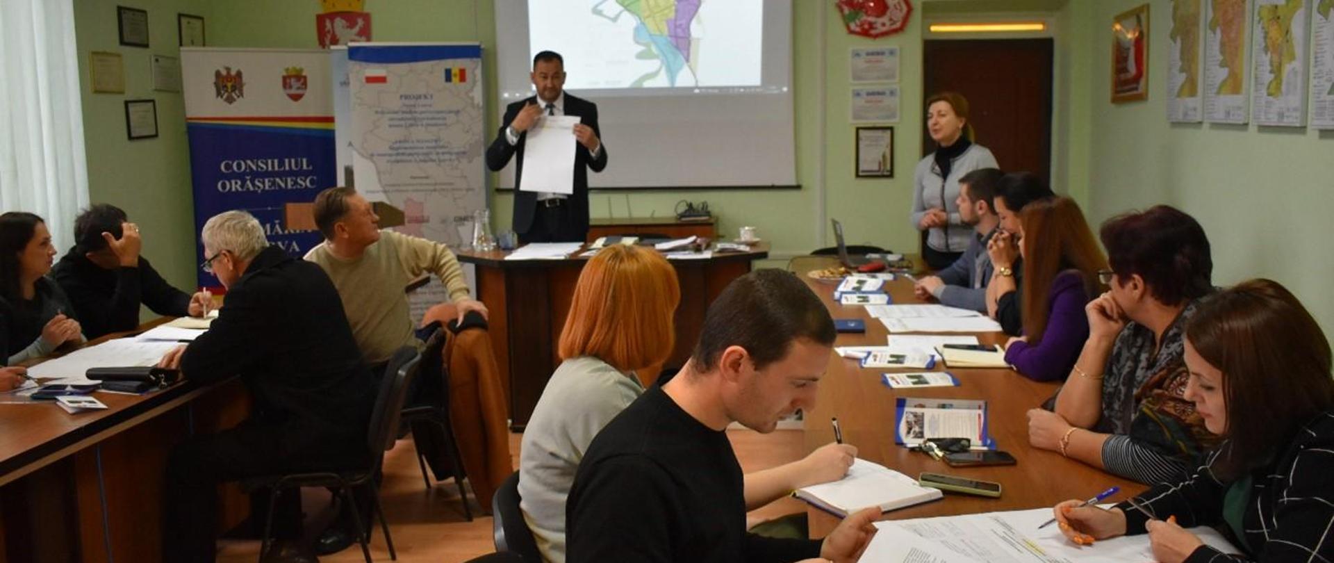 Kilkanaście osób siedzi przy dwóch stołach w trakcie szkolenia, prowadzący pokazuje coś na kartce papieru, W tle dwa rollupy i ekran do prezentacji multimedialnych.