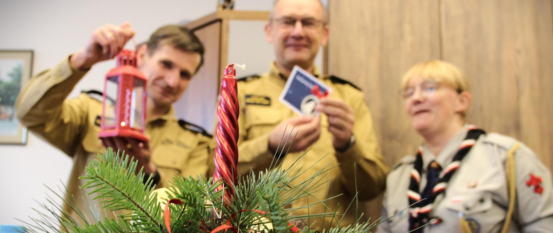 Na pierwszym polanie stroik świąteczny z zielonego świerku z czerwoną świeczką. Za nim stoi dwóch strażaków i harcerka. Prezentują upominki.