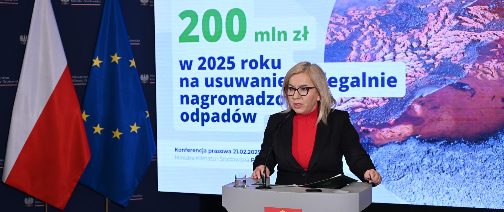 Ministra klimatu i środowiska podczas konferencji prasowej nt. dofinansowania w wysokości 200 mln zł dla samorządów na usuwanie nielegalnie zgromadzonych odpadów. Ministra stoi przy mównicy i mówi do mikrofonu, za nią znajduje się ekran z prezentacją.