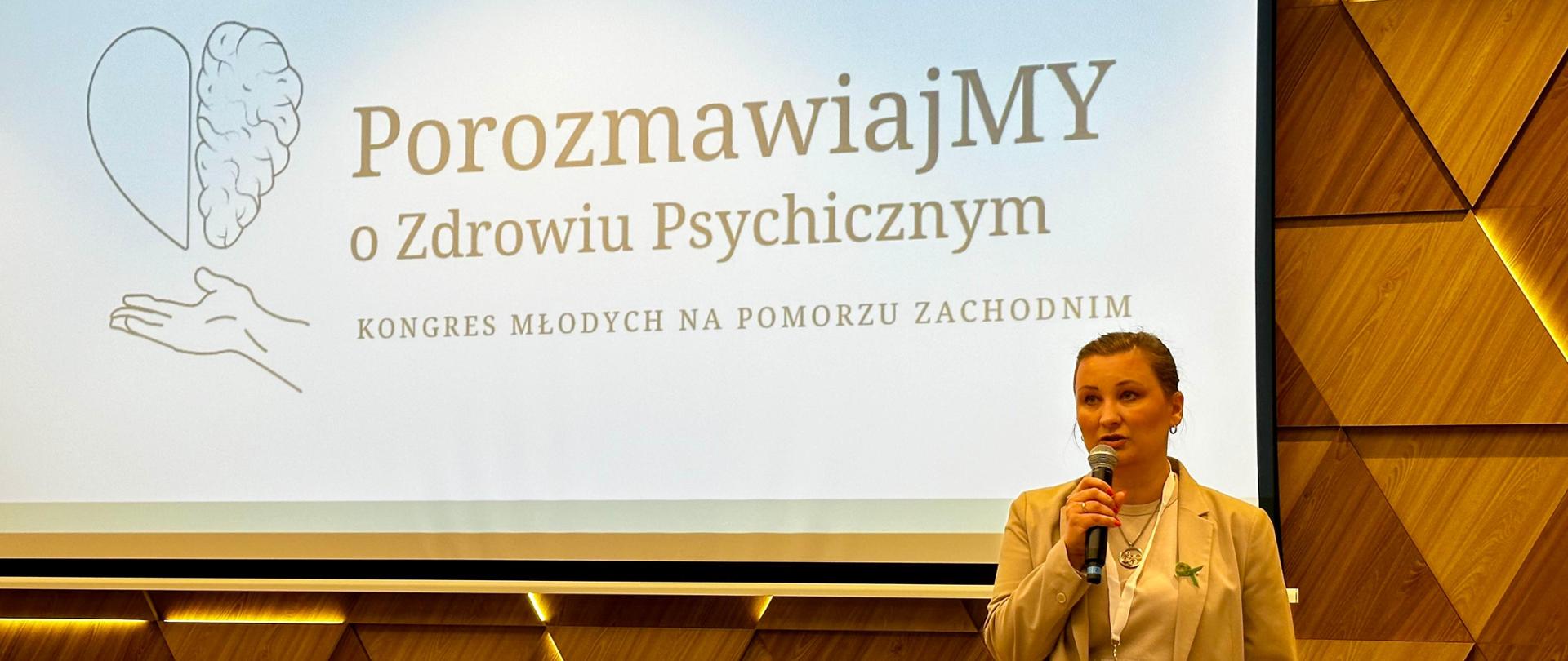 Wiceminister Paulina Piechna-Więckiewicz stoi z mikrofonem w ręku i przemawia. Za jej plecami na drewnianej, podświetlonej ścianie wywieszony jest ekran, na którym wyświetlony jest napis "Porozmawiajmy o Zdrowiu Psychicznym. Kongres Młodych na Pomorzu Zachodnim". 