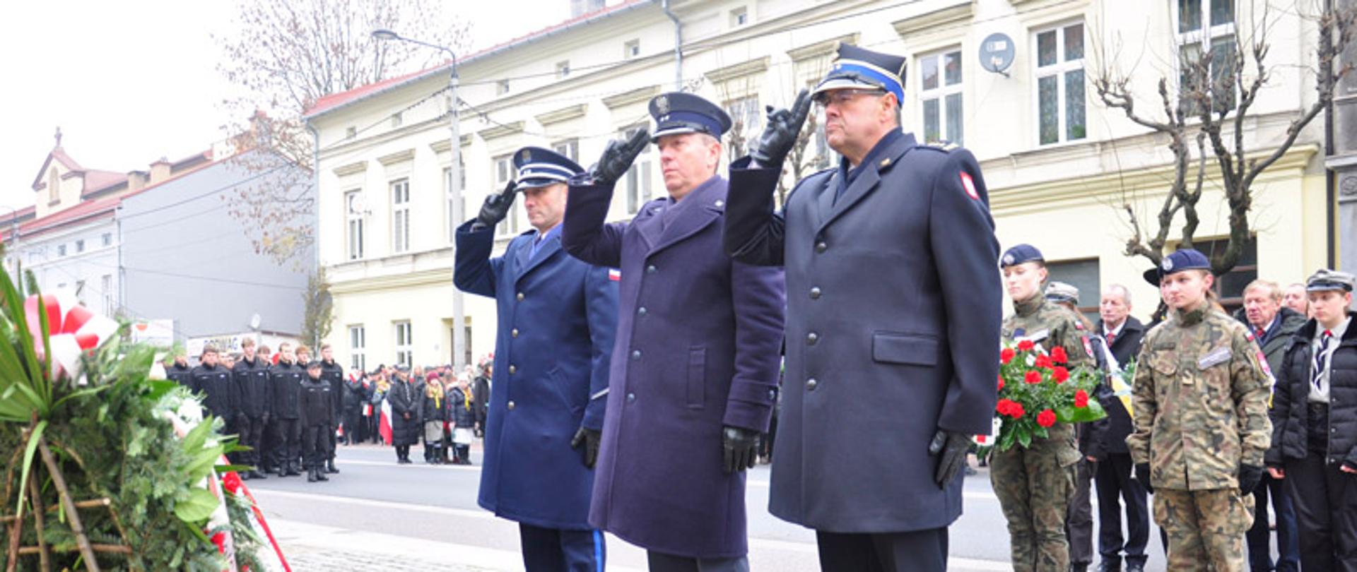 Na pierwszym planie trzech mundurowych, Strażak, Policjant i Strażnik Więzienny, w mundurach wyjściowych oddający honory w trakcie uroczystego składania wieńca