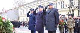 Na pierwszym planie trzech mundurowych, Strażak, Policjant i Strażnik Więzienny, w mundurach wyjściowych oddający honory w trakcie uroczystego składania wieńca