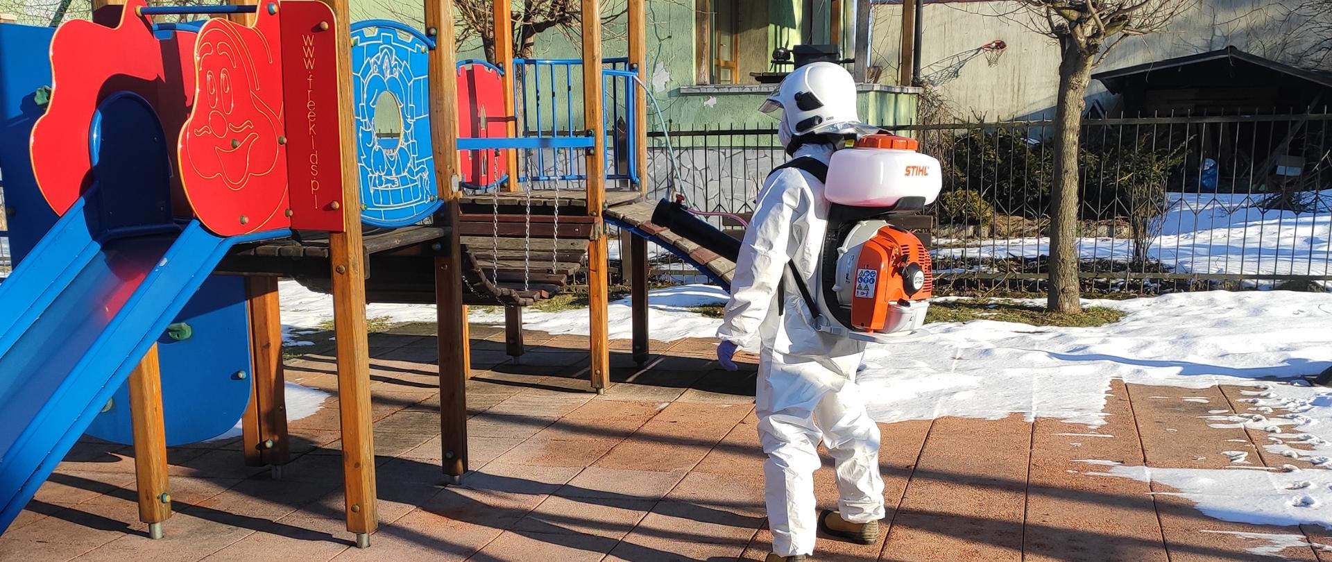 Strażak ubrany w biały struj ochronny przy użyciu opryskiwacza spalinowego dokonuje dezynfekcji zjeżdżalni dla dzieci na placu zabaw