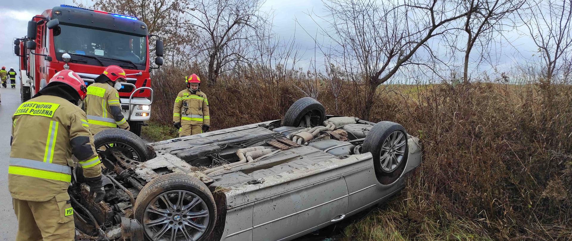 Zdjęcie zrobione w dzień. Zdjęcie przedstawia pojazd, który uległ wypadkowi oraz strażaków, którzy odłączają zasilanie elektryczne w pojeździe. W dalszej części zdjęcia widać samochód strażacki i drzewa. 