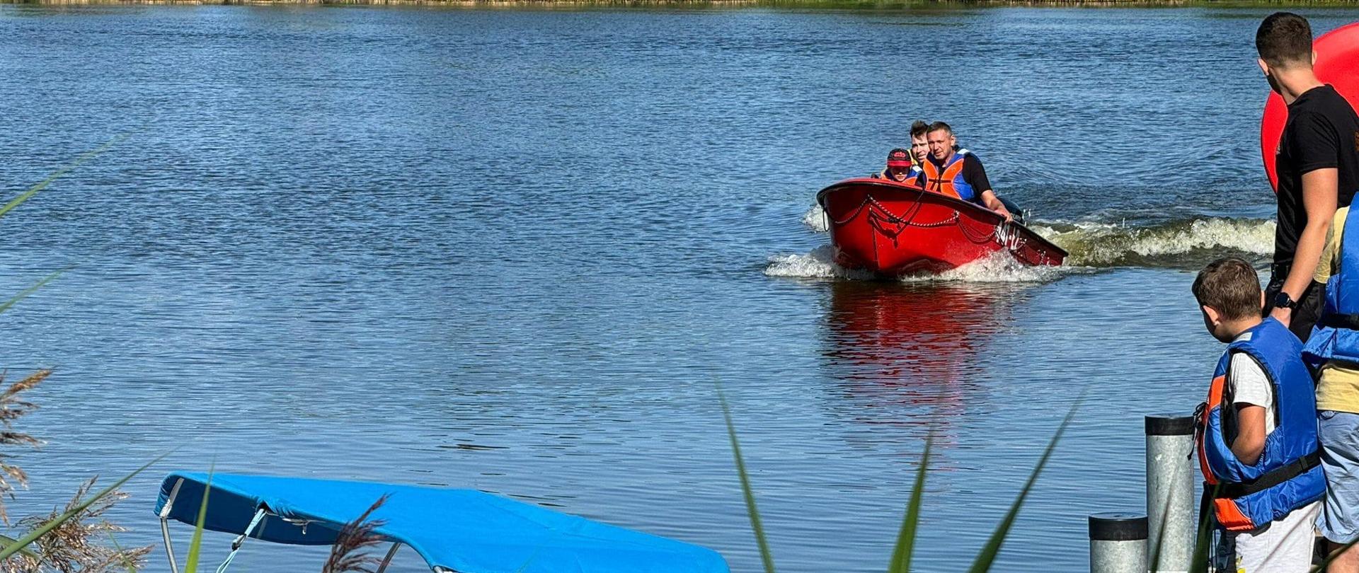 Na zdjęciu widać czerwoną łódkę na wodzie, którą płyną trzy osoby. Na pomoście widać dziecko oraz osobę dorosłą, którzy obserwują osoby pływające. W tle widać bloki, drzewa oraz zarośla. Jest słonecznie.