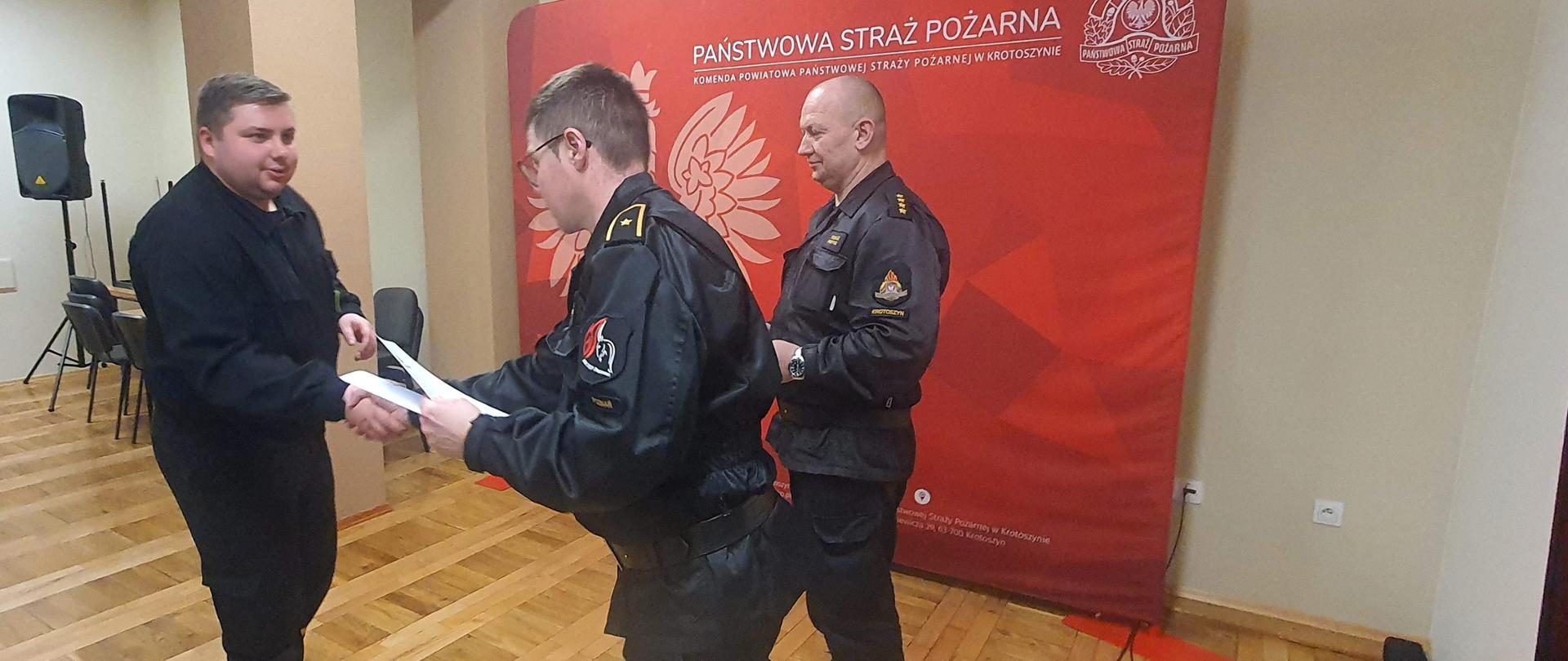 Organizatorzy szkolenia wręczają dyplomy ukończenia szkolenia dla dowdóców OSP. Uroczystość ma miejsce na świetlicy komendy powiatowej. Jeden z członków komisji na tle baneru komendy wręcza dyplom składając gratulację natomiast drugi uczestnik egzaminu przedstawia listę osób, które uzyskały pozytywny wynik.