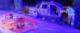 Samochód porozbijany za chodnikiem przy nim strażak z lewej strony na macie koloru pomarańczowego rozłożony sprzęt do ratownictwa drogowego oraz torba medyczna pomiędzy samochodem widoczne drzewka ozdobne