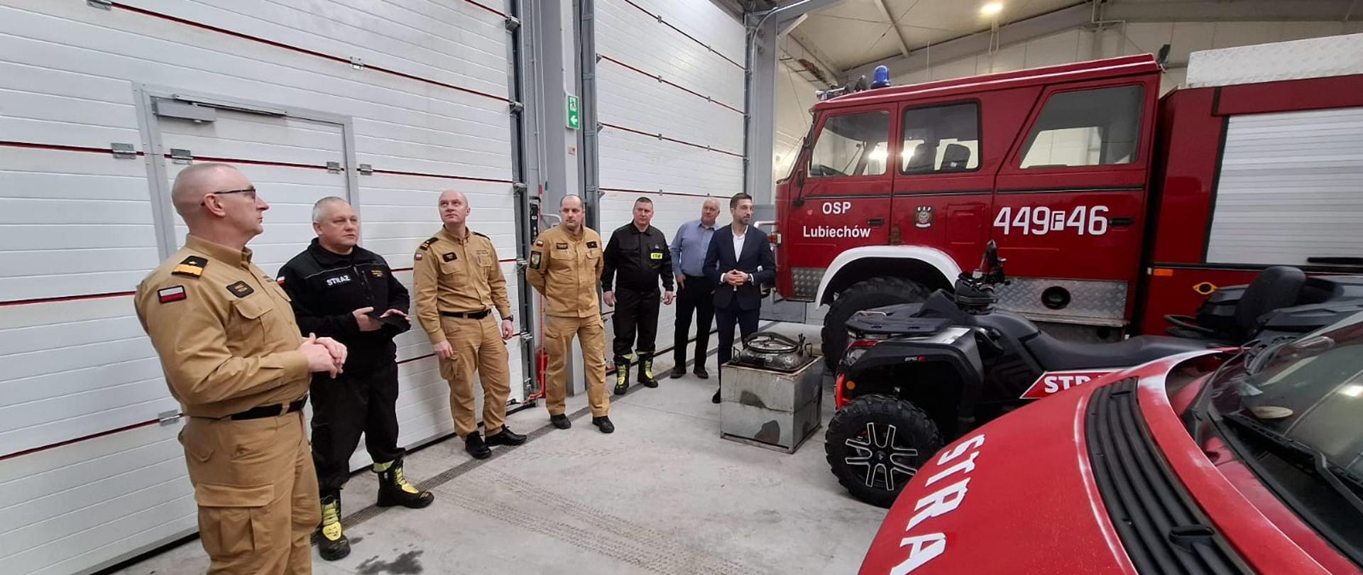 Grupa strażaków w mundurach PSP oraz przedstawiciele OSP stoją w nowoczesnej remizie garażowej. Na pierwszym planie widoczne są wóz strażacki oraz quad z oznaczeniami straży pożarnej.