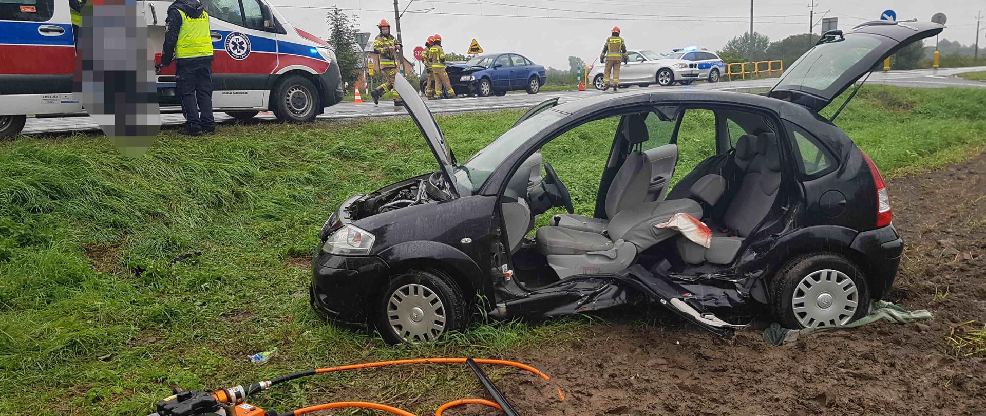 Na pierwszym planie uszkodzony samochód osobowy znajdujący się poza jezdnią oraz sprzęt hydrauliczny używany do wykonania dostępu do osoby poszkodowanej. Na drugim planie ambulans, ratownicy medyczni, policjanci i strażacy prowadzący czynności na miejscu zdarzenia.