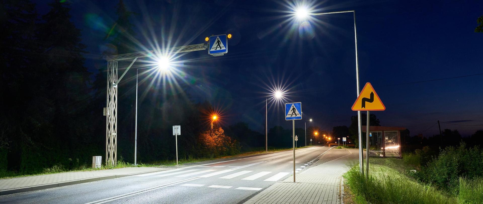 Widok z poziomu ziemi na oświetlone przejście dla pieszych. Pora nocna, widać oświetlenie drogi w tym to nad przejściem dla pieszych. Z prawej strony jest chodnik, który prowadzi do przystanku autobusowego. 