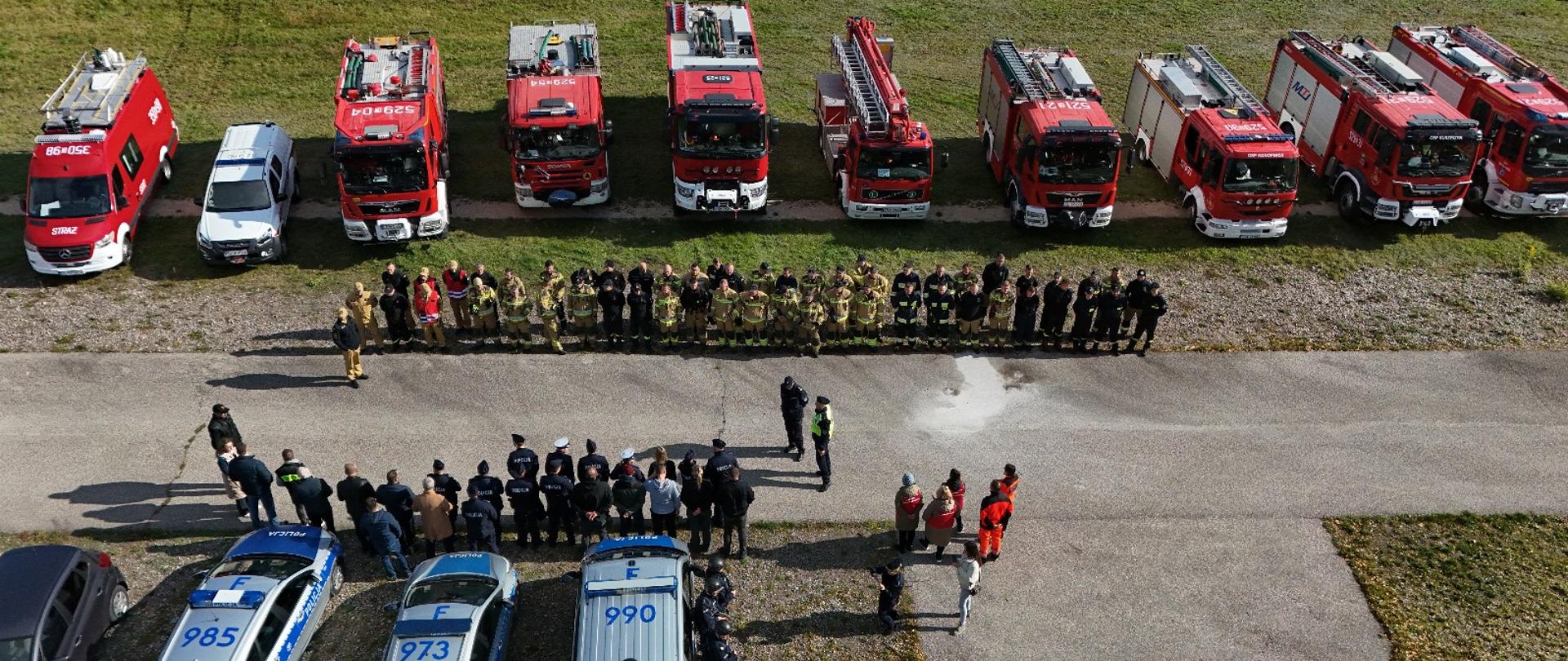 Na zdjęciu po środku znajduje się droga, z jednej strony drogi w rzędzie ustawionych jest 10 samochodów strażackich, a przed nimi stoją strażacy i druhowie OSP. Natomiast po drugiej stronie drogi stoją 3 samochody policyjne oraz policjanci. Zdjęcie zrobione jest z góry.