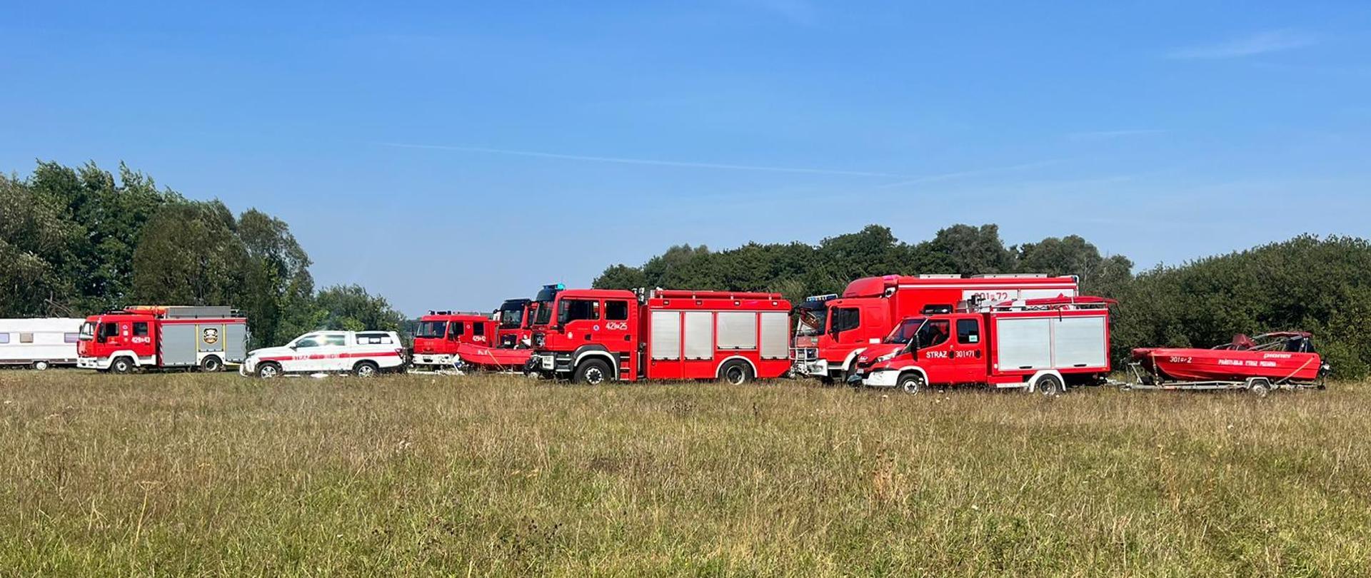 Na zdjęciu widać samochody pożarnicze, które zostały zadysponowane do zdarzenia. Pojazdy stoją na łące bezpośrednio przyległej do jeziora. W miejscu tym wodowano łodzie ratownicze.