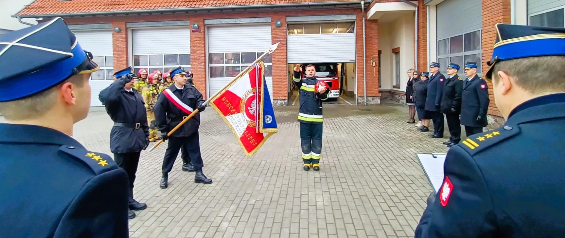 Zdjęcie przedstawia uroczysty moment składania ślubowania przez nowo przyjętego strażaka Mariusza Kłoczko do służby w Komendzie Powiatowej Państwowej Straży Pożarnej w Gołdapi. Z lewej strony obrazu znajduje się część postaci dowódcy jednostki ratowniczo-gaśniczej kpt. Adriana Korotko, natomiast z prawej strony część postaci Komendanta Powiatowego Państwowej Straży Pożarnej w Gołdapi st. bryg. Wojciecha Szczepanika, który przyjmuje ślubowanie. W tle można zaobserwować pozostałych strażaków biorących udział w uroczystej zbiórce oraz pojazdy pożarnicze zlokalizowane w garażu strażnicy