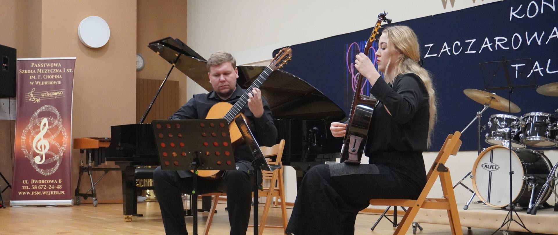 Na scenie duet gitarowy gra w składzie Piotr Kłosowski i Zofia Marszk. W tle widać fortepian oraz instrumenty perkusyjne. Muzycy grają na siedząco patrząc w pulpit.