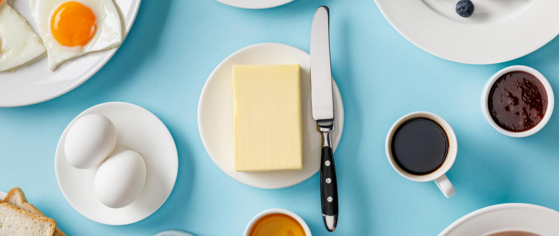 Na zdjęciu w centrum na talerzyku znajduje się masło a obok nóż. Wokół są jajka, tosty, borówki i kawa. Tło jest niebieskie.