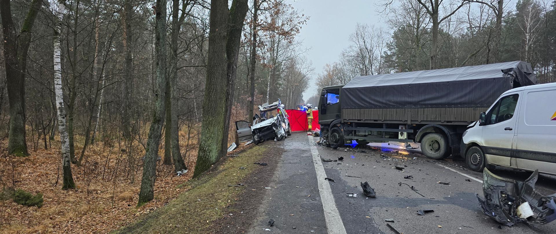 Na zdjęciu widoczny jest wrak samochodu osobowego pocięty przez strażaków sprzętem hydraulicznym. Na dalszym planie widać czerwony parawana z napisem Straż. Widoczni są również strażacy i żołnierze oraz wojskowa ciężarówka i biały bus.