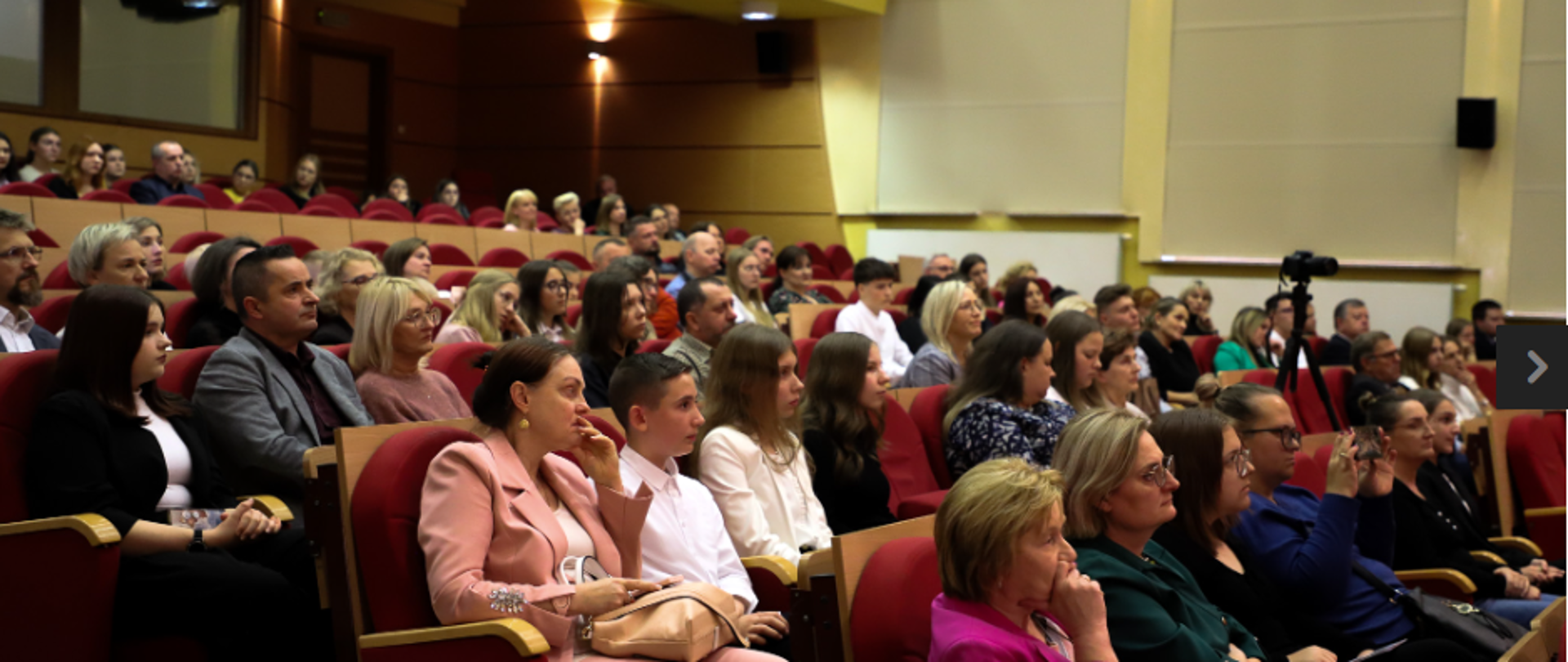 Sala- aula konferencyjna i wiele osób siedzących. Światła na ścianach - jest bardzo jasno.