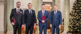 W szeregu stoją minister Kulasek, prezydent Duda, były minister Wieczorek i premier Tusk, za nimi ściana z marmurowymi kolumnami, z prawej pod ścianą ozdobiona bombkami i łańcuchami choinka.