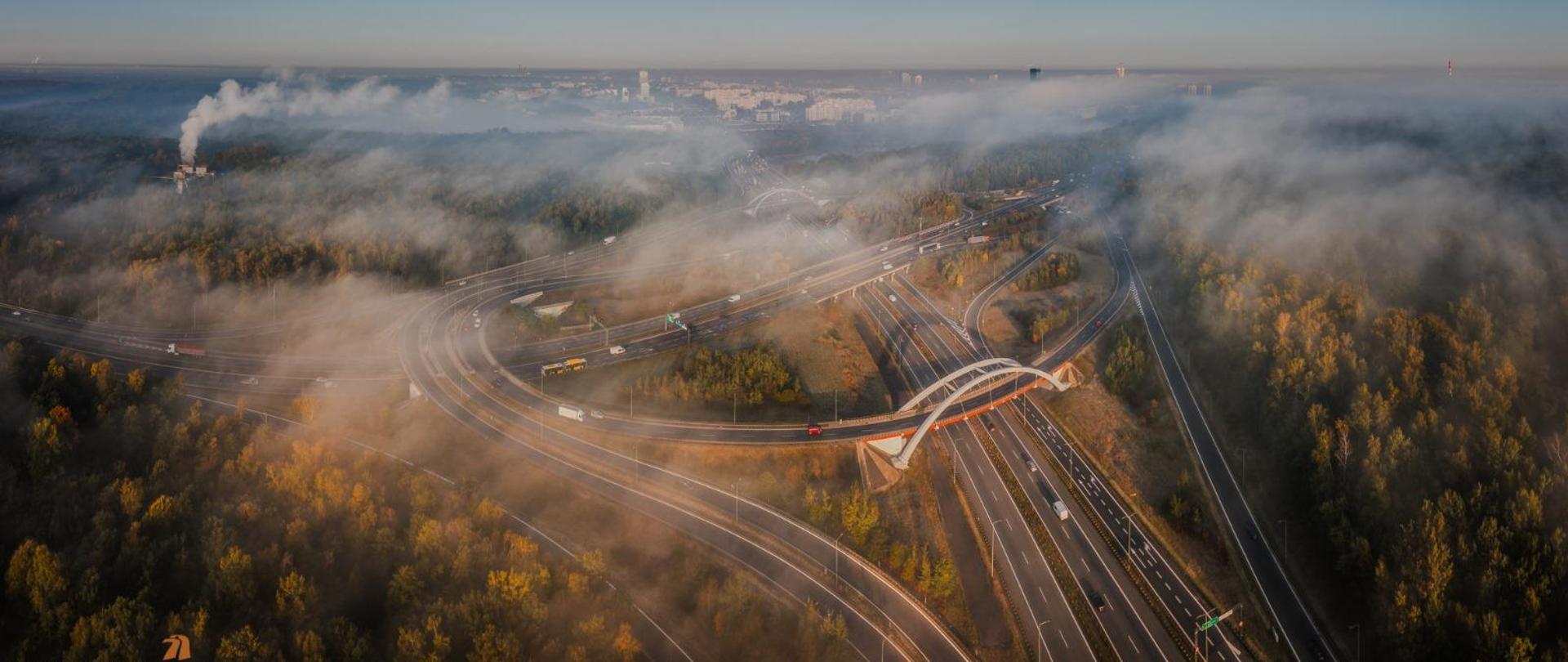 Zdjęcie lotnicze węzła na autostradzie A4. Zachodzące słońce, droga spowita mgłą przebiega przez las.