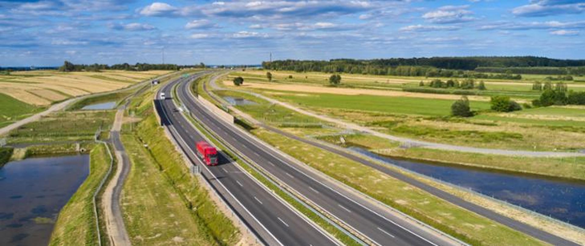 Widok z góry na drogę ekspresową S19. Pogoda słoneczna, zielona trawa po obu stronach drogi. Droga ekspresowa biegnie po łuku, jadą po niej auta ciężarowe. Na pierwszym planie czerwony tir. Po obu stronach drogi widać zbiorniki retencyjne.