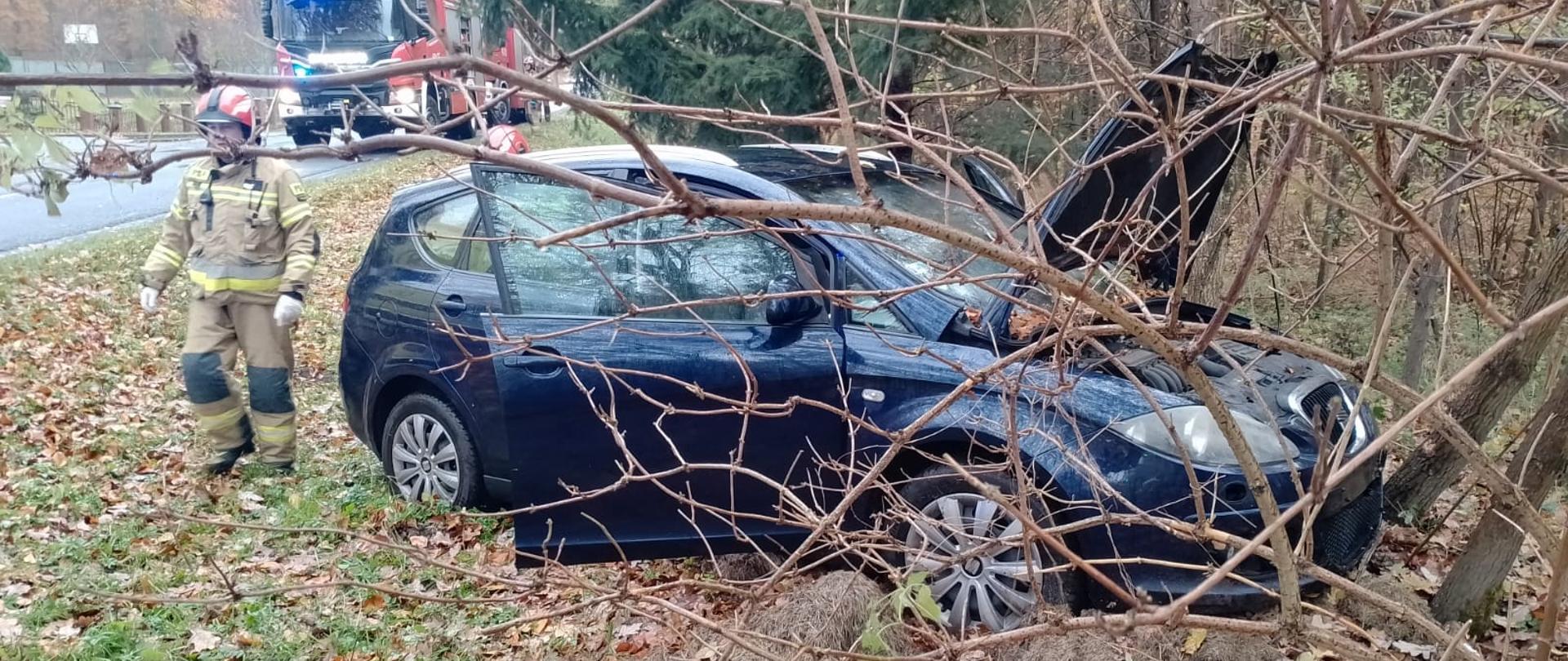 Samochód osobowy między drzewami z podniesioną maską, obok strażak, w tle samochód pożarniczy