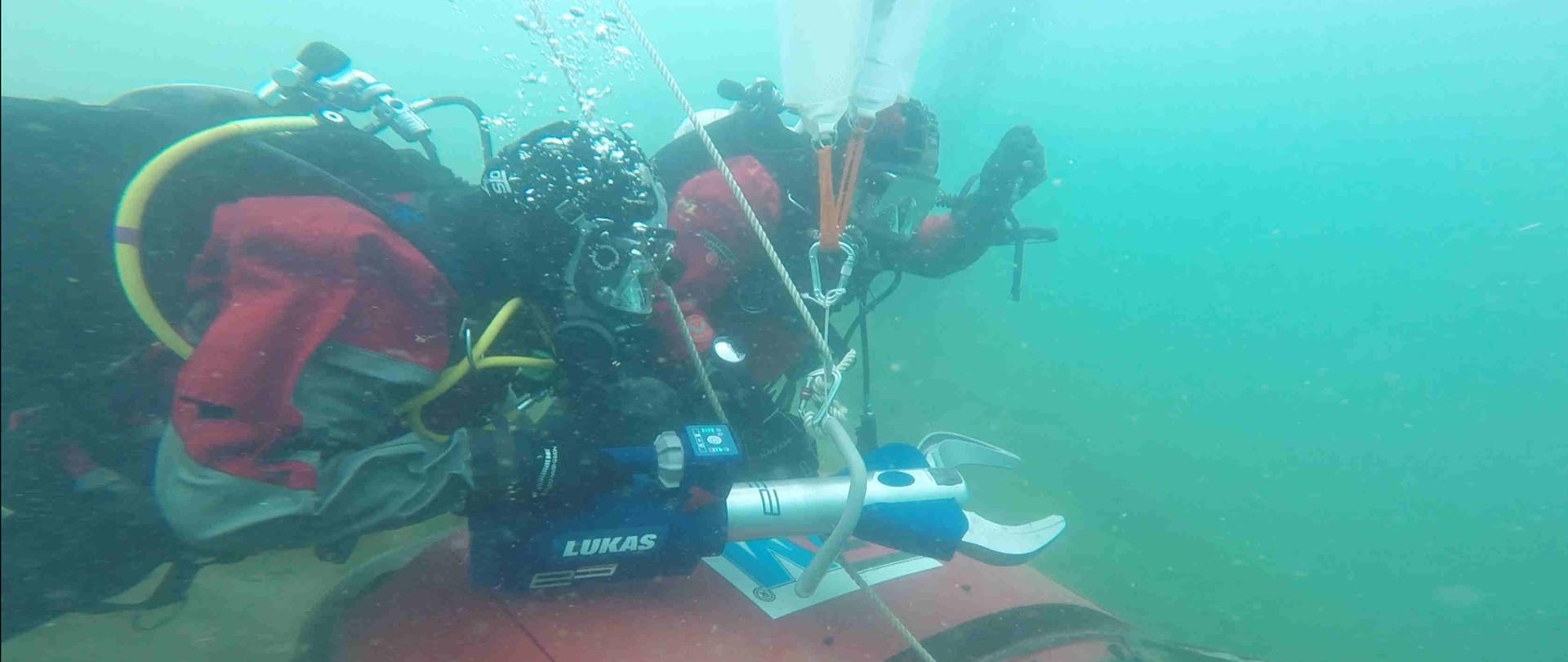 Zdjęcie zrobione pod wodą. Na zdjęciu widać dwóch strażaków – nurków, którzy sprzęt hydraulicznym pracują pod wodą, widać samochód, który tną. 