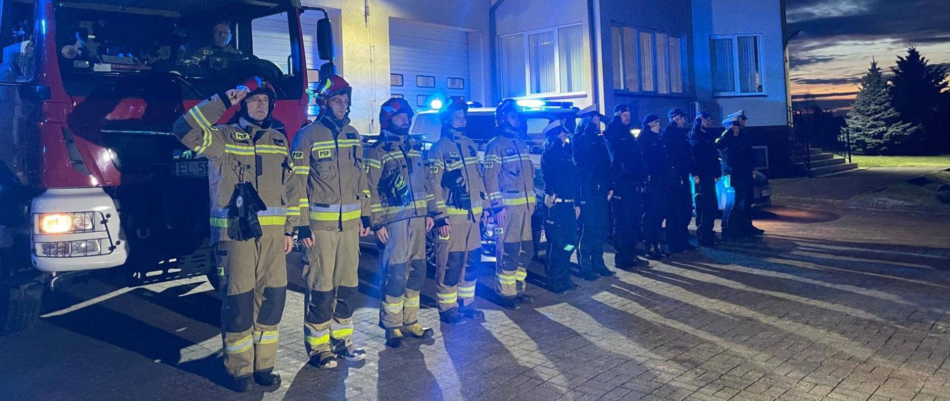 Przed budynkiem komendy państwowej straży pożarnej w jednym rzędzie stoi kolejno 5 strażaków i 8 policjantów. Za funkcjonariuszami stoi samochód bojowy strażacki oraz policyjny samochód osobowy z włączonymi sygnałami świetlno-dźwiękowymi.