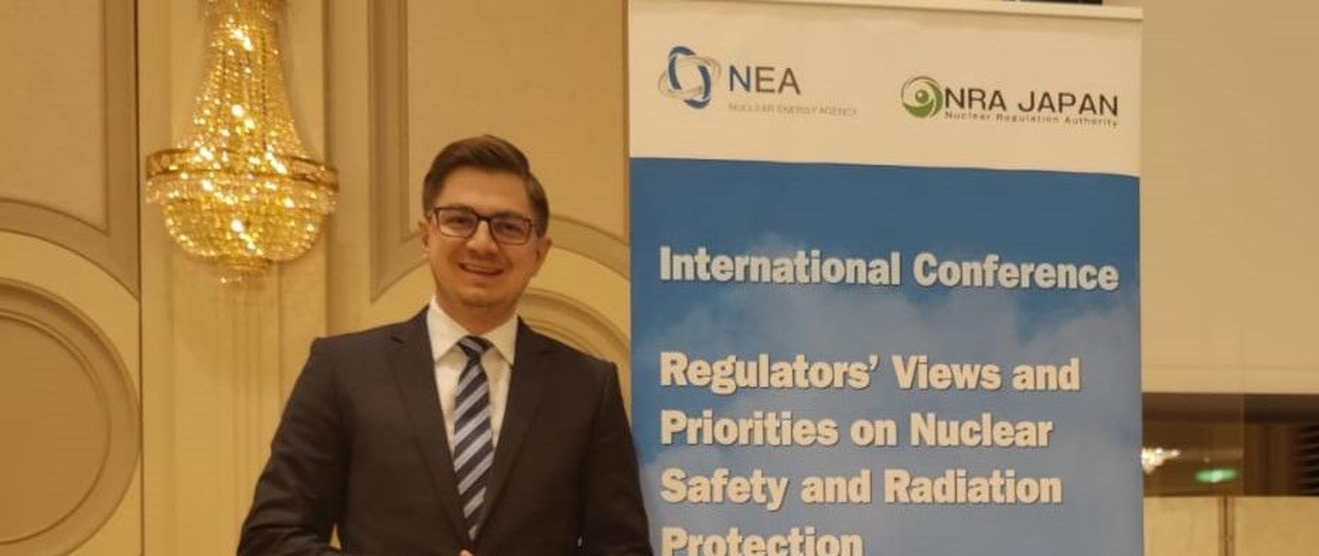 The President of the PAA at the conference on priorities in nuclear safety - Dr. Łukasz Młynarkiewicz is standing next to the banner promoting the conference