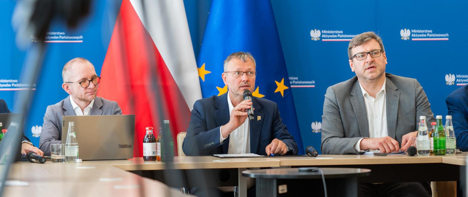 Wiceminister Jacek Bartmiński siedzi w garniturze przy stole. W ręku trzyma mikrofon. W tle ścianka i flagi. Po obu stronach siedzą pracownicy resortu. 