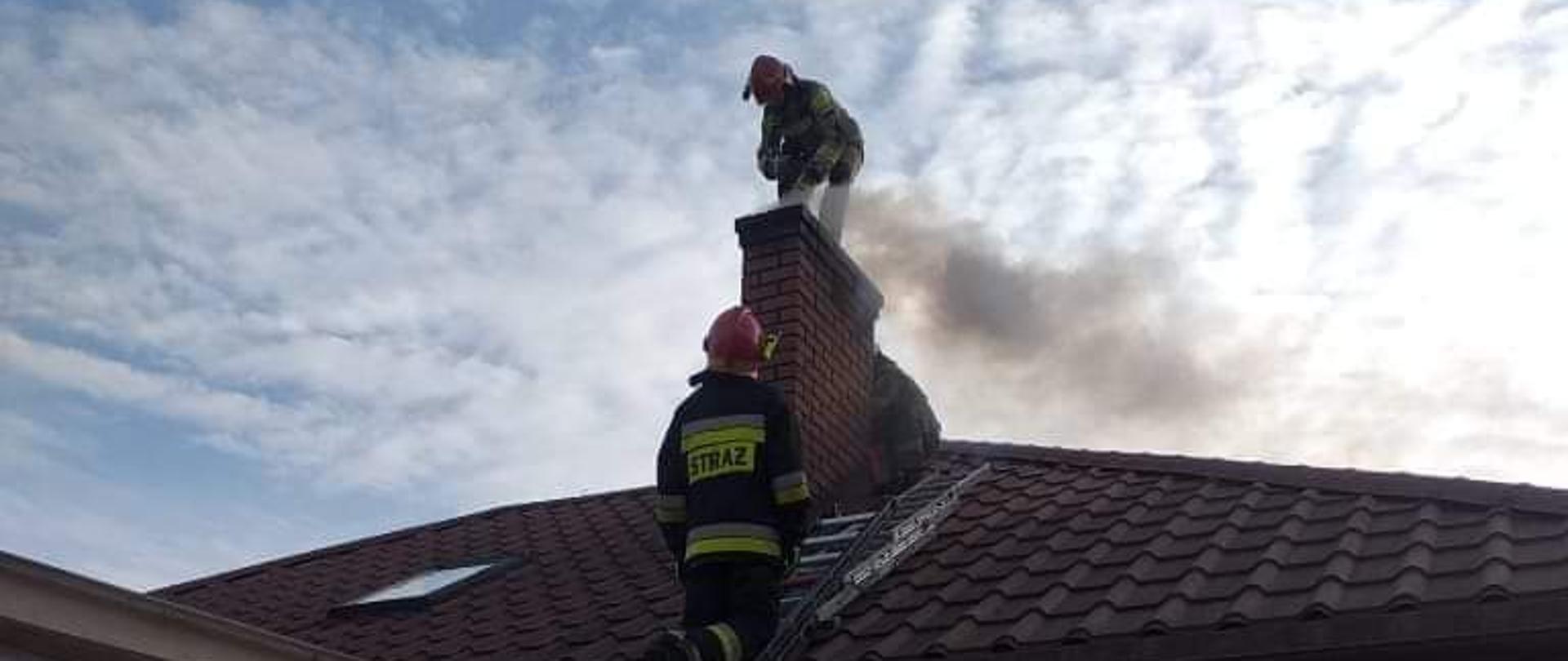 Na dachu domu stoją dwaj strażacy, którzy gaszą palące się sadze w kominie. W tle błękitne niebo. 
