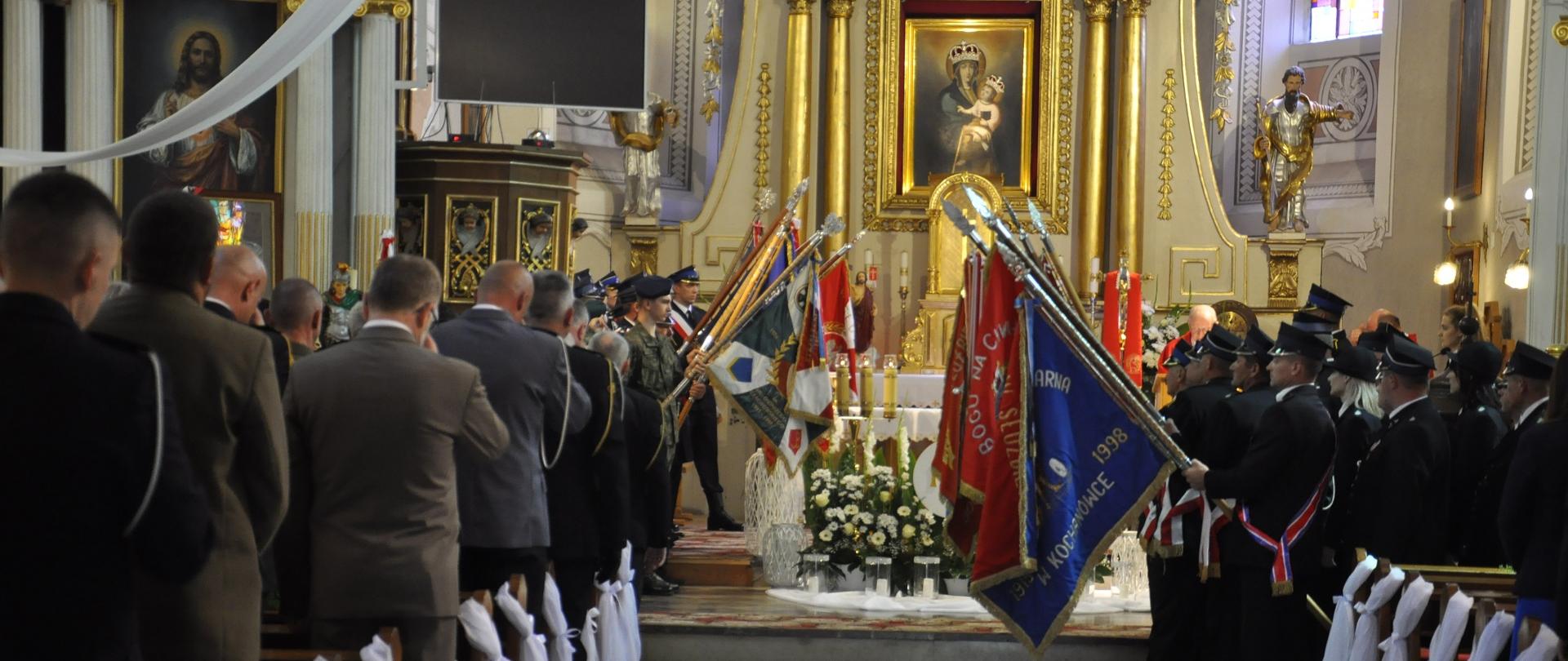 Zdjęcie zrobione z końca kościoła.Widać na nim oddające honor sztandary pochylone w stronę ziemi oraz gości stojących w ławkach kościelnych.