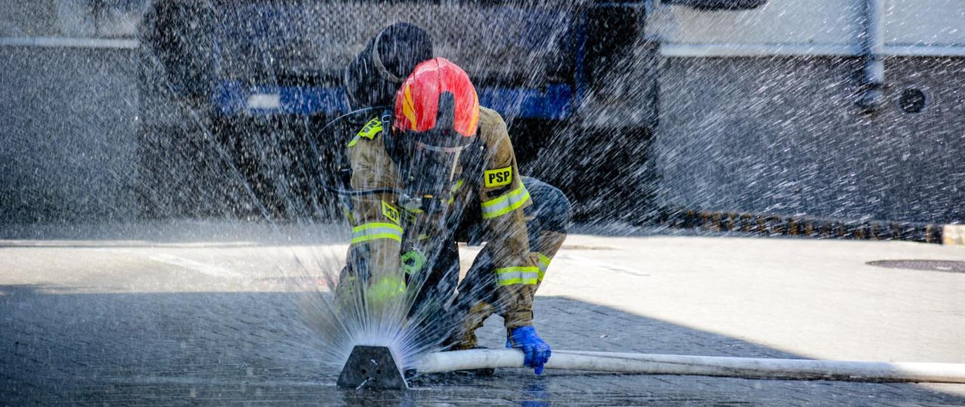 strażak w pełnym umundurowaniu specjalnym klęczący na prawnym kolanie w trackie wytwarzani zasłony wodnej 