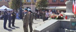Moment oddawania honoru przez 3-osobową delegację mężczyzn w mundurach przed pomnikiem, pod którym złożone są wiązanki kwiatów.
