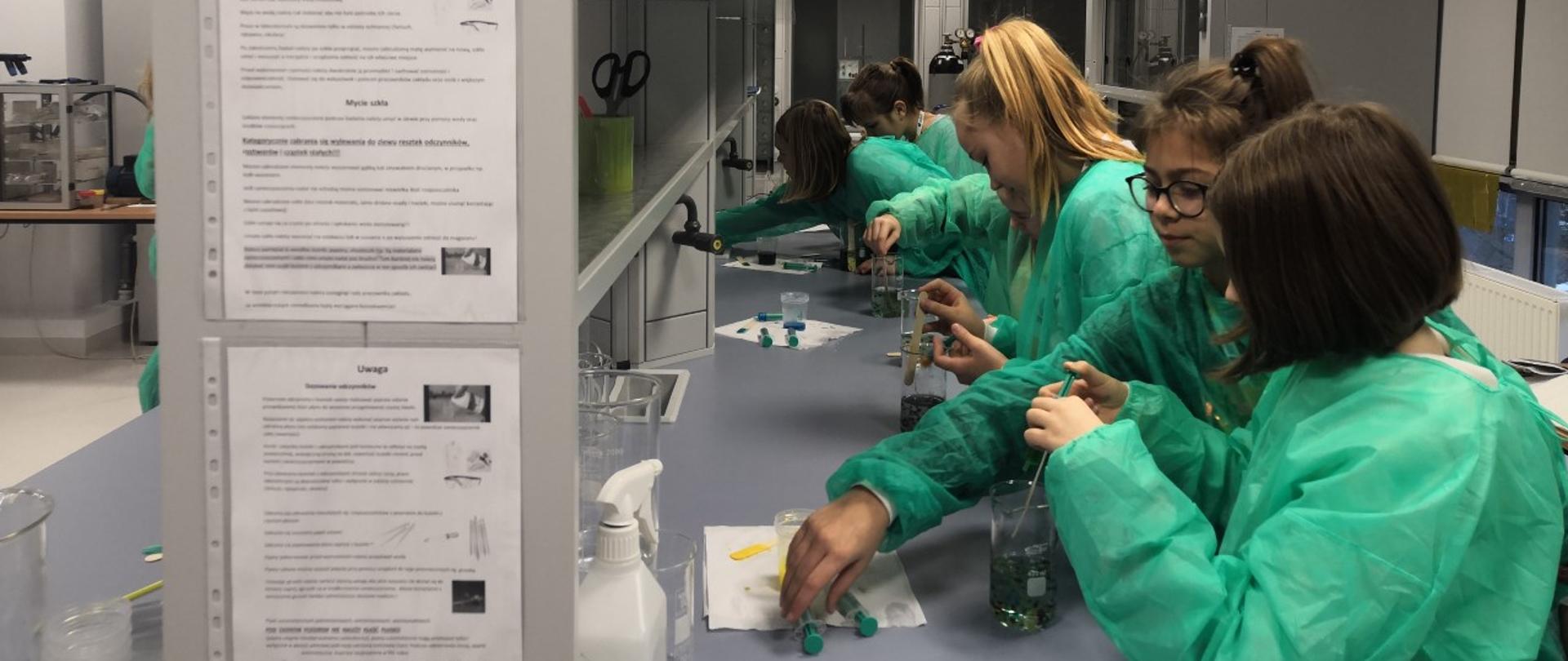 Classes in the laboratory. Seven teenagers in green aprons, standing in a row by the long table top, pour the liquid into the measuring cups using syringes.