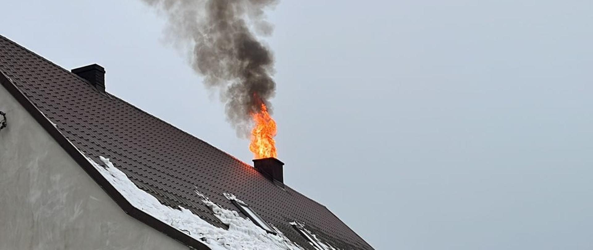 Dom jednorodzinny. W komina wydobywa się ogień oraz kłęby dymu. Przed domem widoczny fragment ogrodzenia.