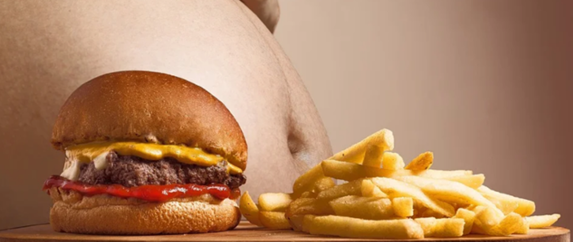 Na zdjęciu po lewej stronie widoczny jest burger, natomiast po prawej frytki. W tle widoczny jest brzuch osoby otyłej.