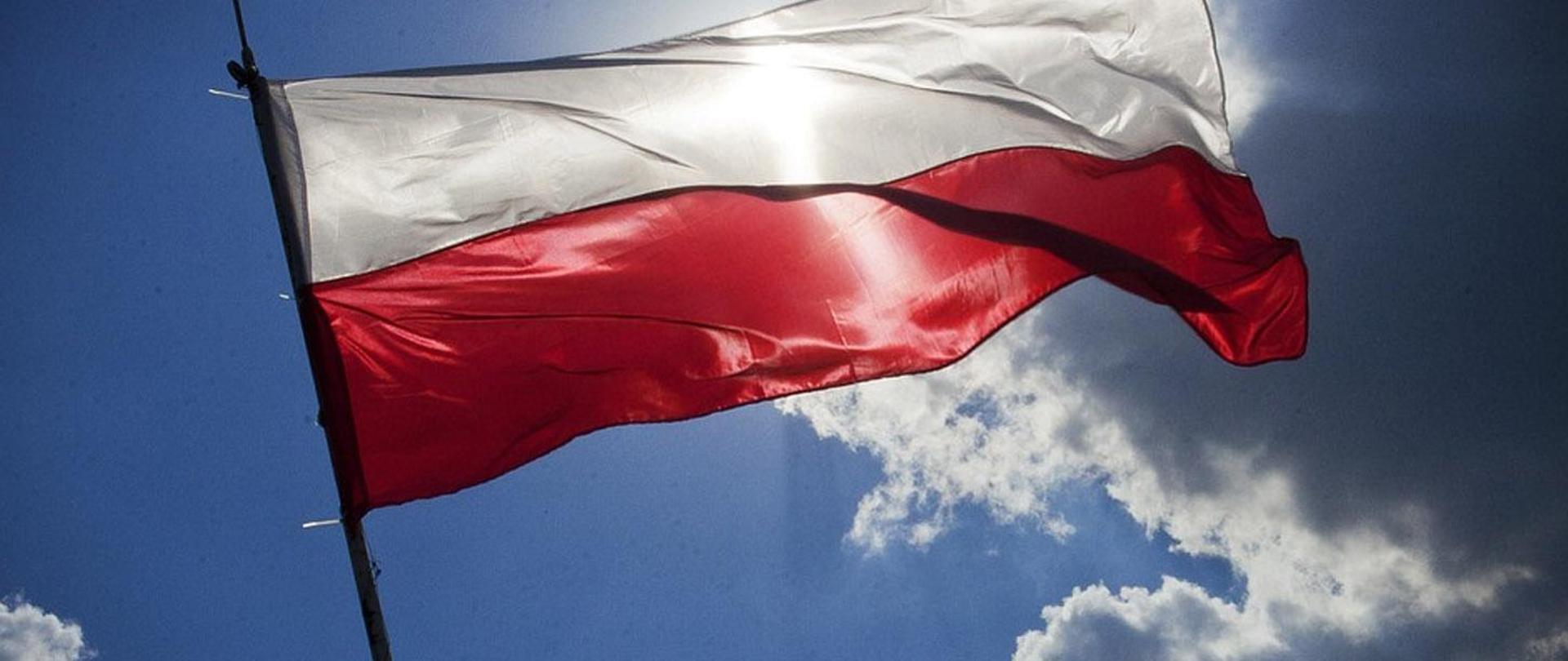 Na zdjęciu znajduje się biało - czerwona państwowa flaga Rzeczypospolitej Polskiej. Flaga znajduje się na tle błękitnego nieba.