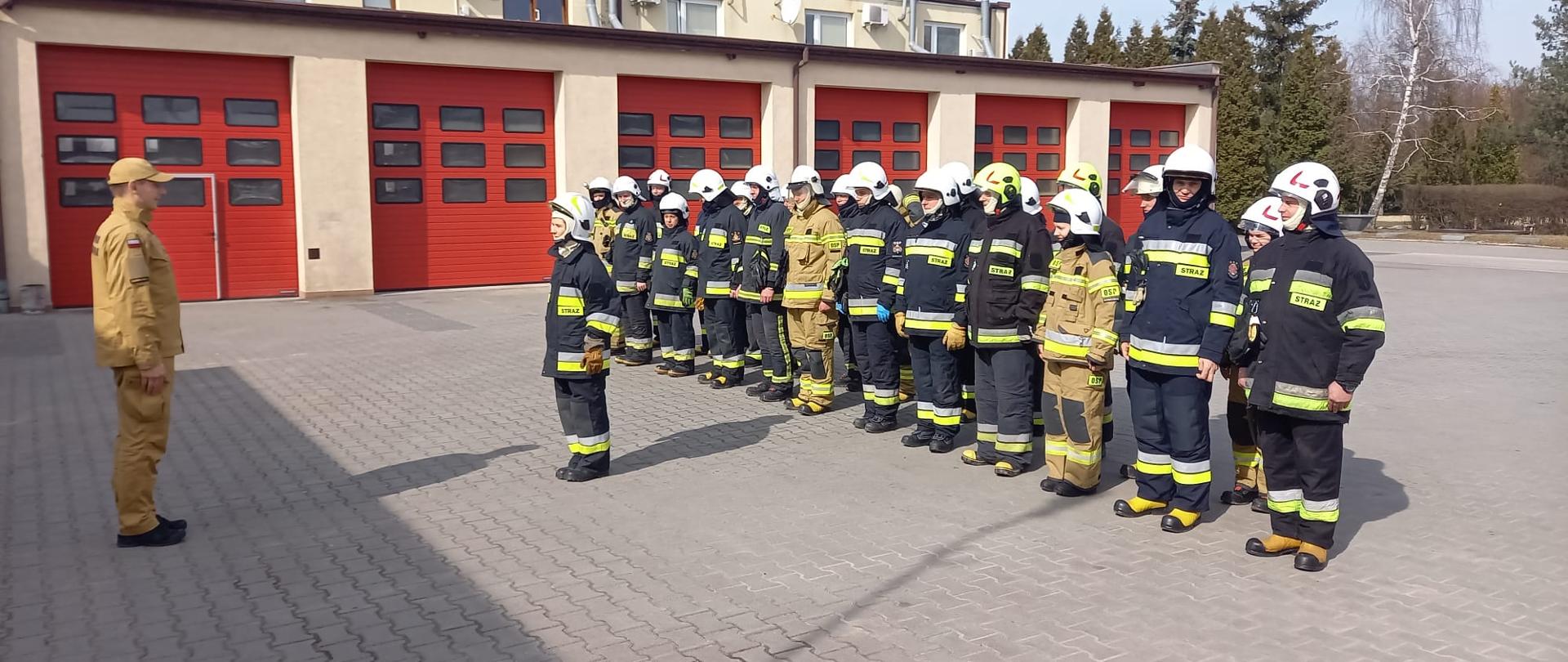 Zdjęcie przedstawia zbiórkę strażaków OSP przed garażami JRG w Zduńskiej Woli. Funkcjonariusz z PSP przyjmuje meldunek od Druhny z OSP o gotowości ratowników do egzaminu. 