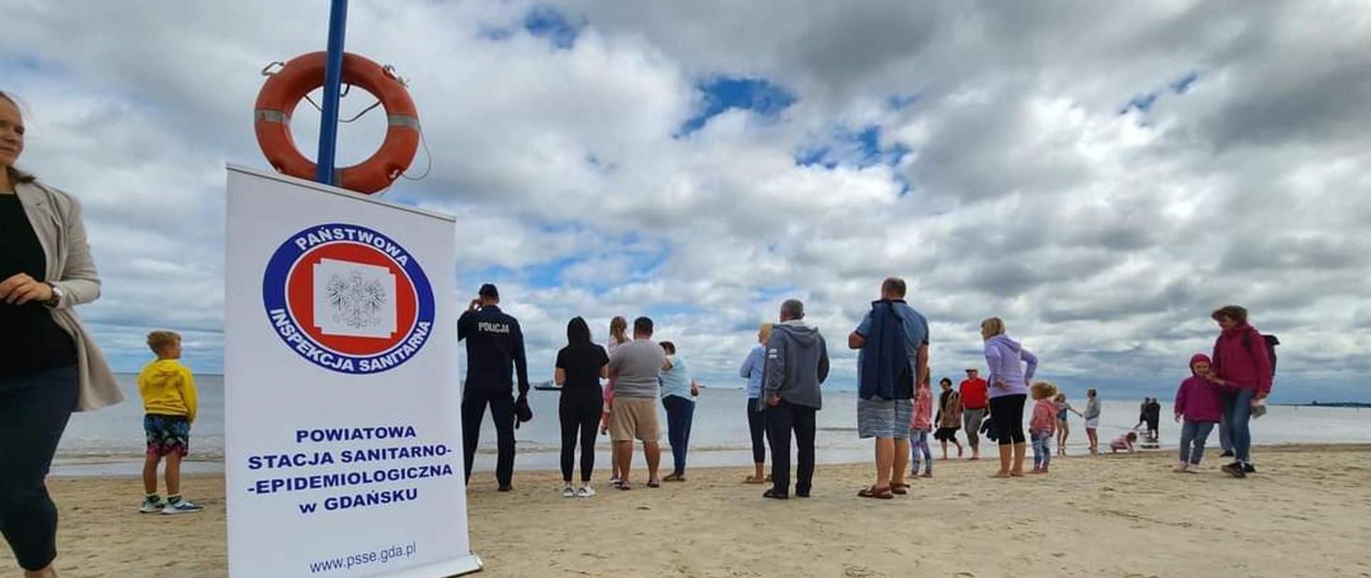 Policjant oraz grupa osób stojących na plaży, zwróconych w kierunku morza. Na pisaku stoi roll-up z logo Państwowej Inspekcji Sanitarnej nad którym wisi pomarańczowe koło ratunkowe.