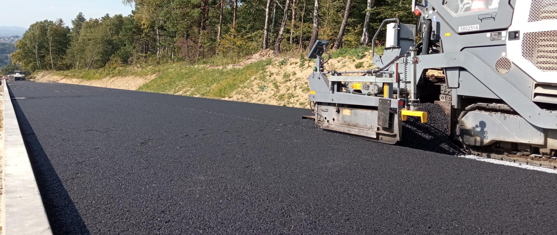 Widok na odcinek próbny nawierzchni wykonywany na budowie drogi ekspresowej S19 Babica - Jawornik. Po prawej stronie widać rozściełacz asfaltu. Świeci słońce, po prawej stronie widać drzewa.