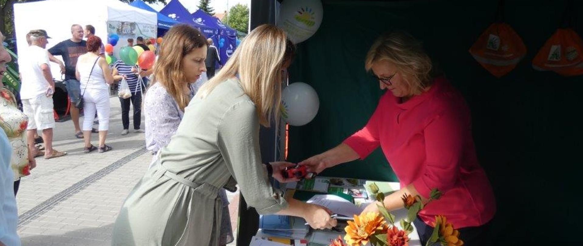 Kobieta i dziewczynka przy stoisku KRUS podaje wypełniony formularz konkursowy kobiecie obsługującej stoisko 