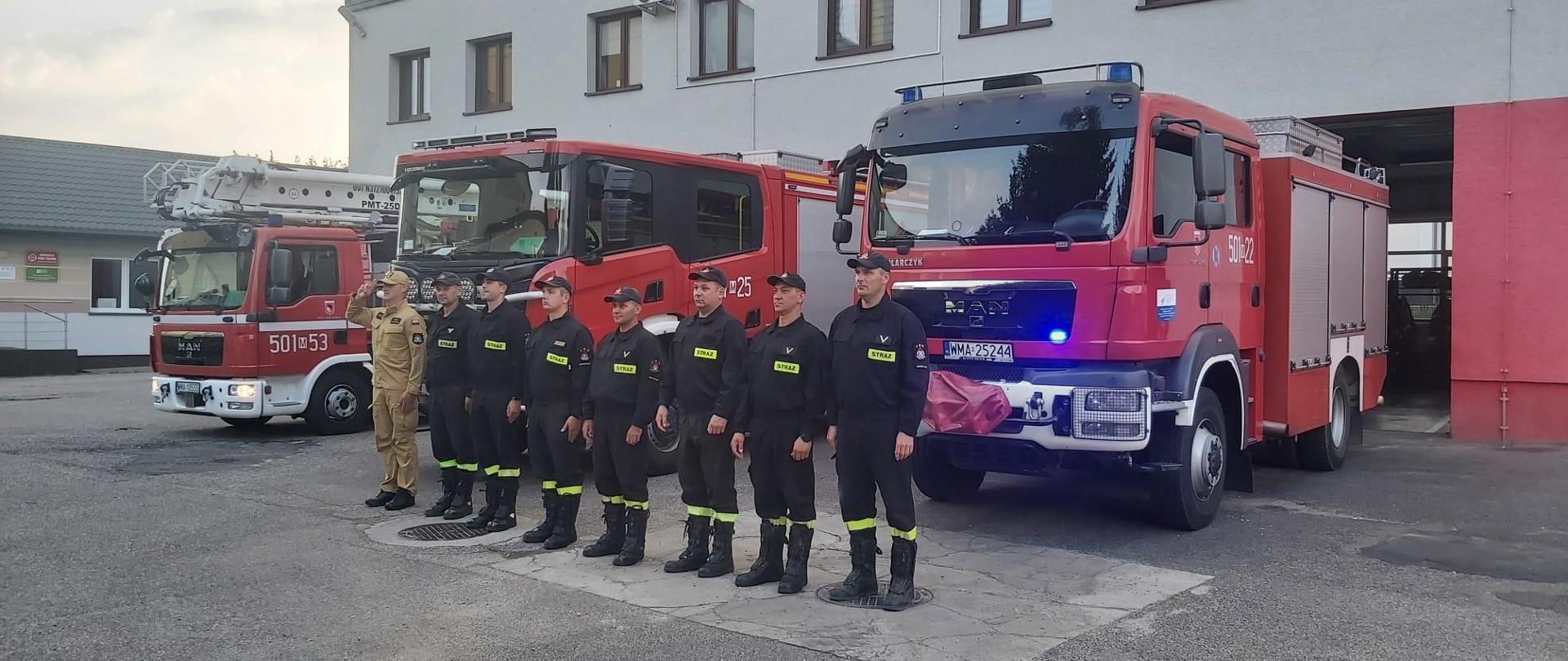 Strażacy stoją w szeregu przed samochodami strażackimi z włączonymi sygnałami świetlnymi i dźwiękowymi. Piwerszy strażak salutuje.