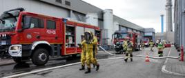 Zdjęcie przedstawia strażaków w trakcie działań ratowniczo-gaśniczych w trakcie ćwiczeń w Zakładach Mięsnych Sokołów S.A. w Jarosławiu. W tle widoczne pojazdy pożarnicze oraz budynki zakładowe.