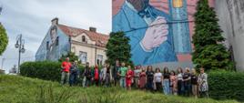 Słuchacze i nauczyciele stojący na tle ścian budynków na których są namalowane postacie Ignacego Łukasiewicza i Jana Szczepanika. 