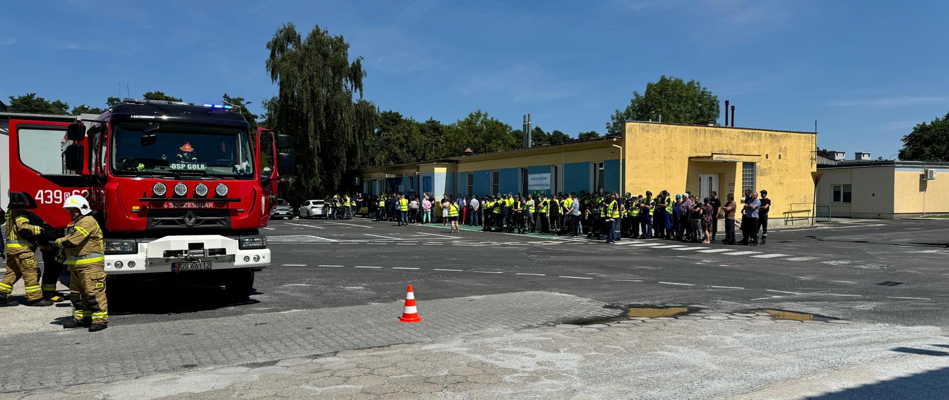 Z lewej strony zdjęcia stoi pojazd pożarniczy. W prawej części stoją wszyscy pracownicy zakładu, którzy zostali ewakuowani z budynku.