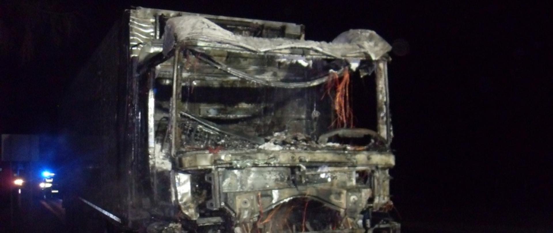 Zdjęcie przedstawia całkowicie spaloną kabinę ciągnika siodłowego, za naczepą widać światła błyskowe w kolorze niebieskim pojazdu służb ratowniczych 