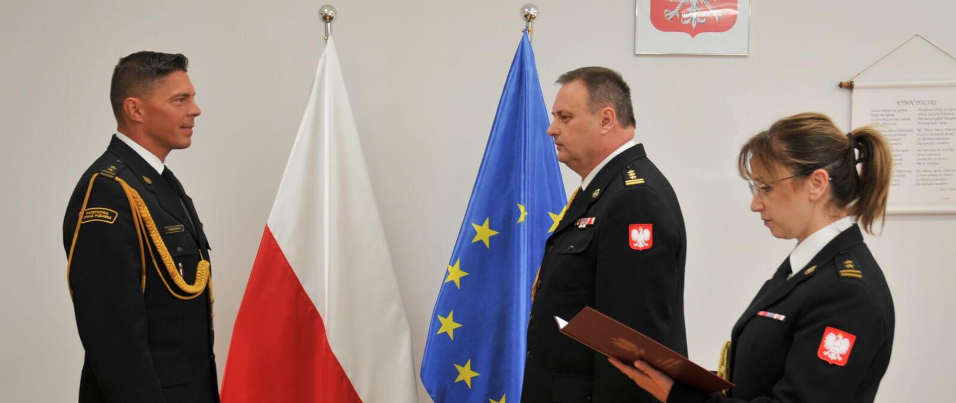 po lewej stoi jednej strażak na przeciwko niego dwoje strażaków, funkcjonariuszka po najbardziej z prawej trzyma w rękach teczkę i odczytuje pismo, za nimi flagi polski i unii europejskiej