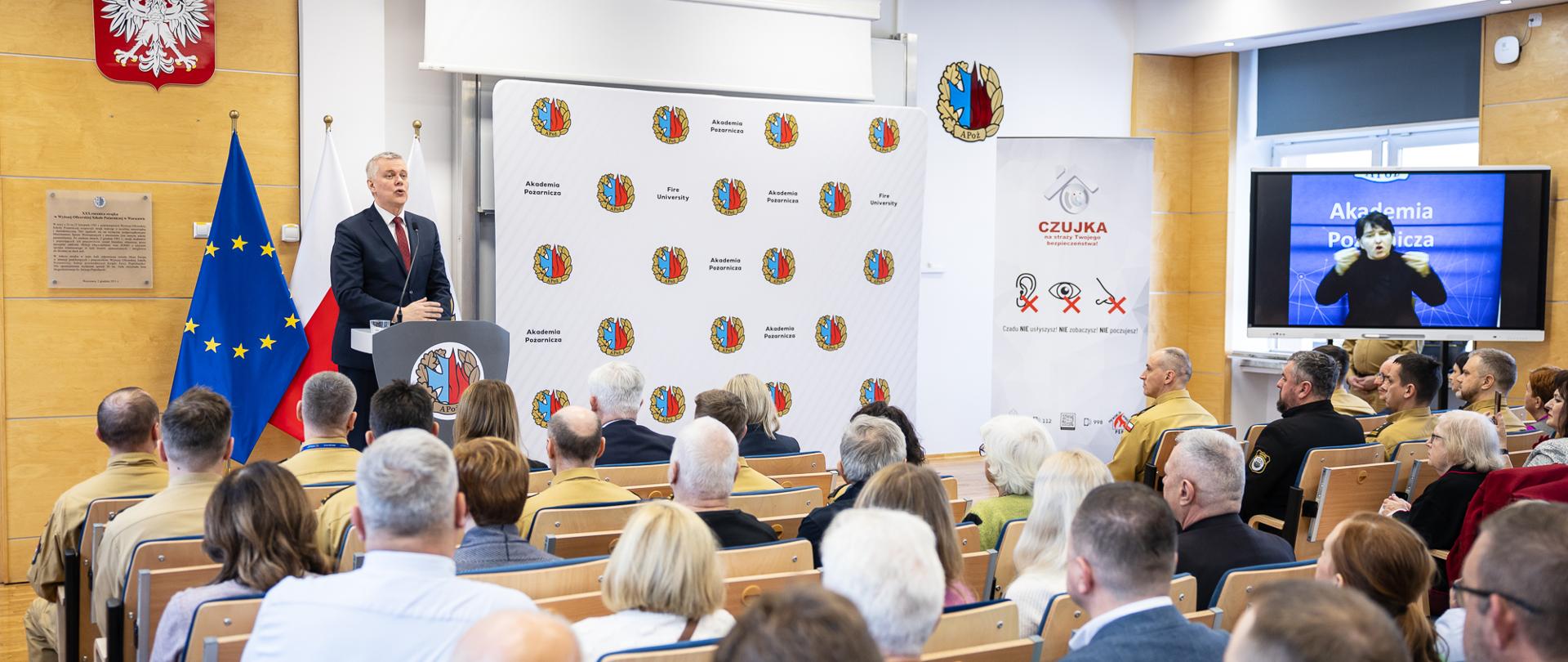 Mężczyzna na tle flag Polski i UE przemawia do siedzących, zwróconych w jego stronę ludzi, uczestników konferencji.