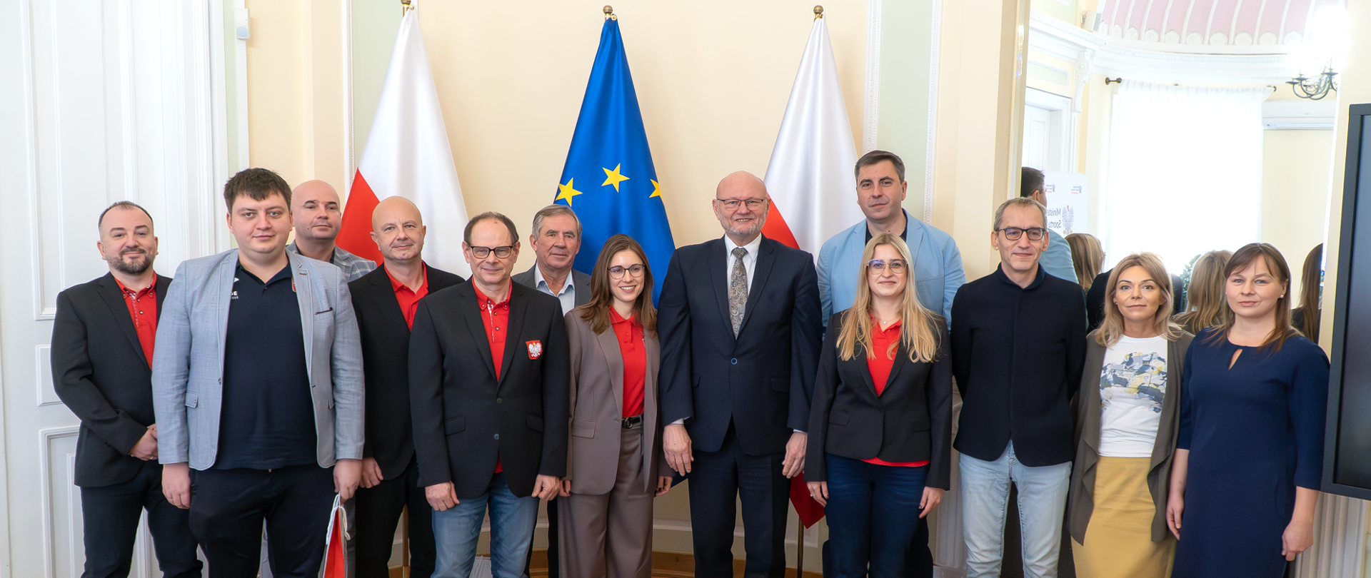 Zbiorowe zdjęcie Ministra Ireneusza Nalazka z reprezentantami polski na 16. World Bridge Games w brydżu sportowym. Za ich plecami dwie flagi Polski, a między nimi flaga UE.